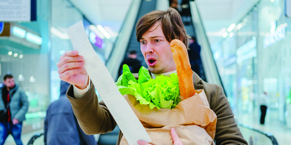 Man on an escalator holding a paper bag of groceries looking a supermarket receipt with a shocked look on his face.