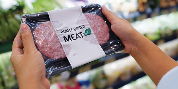 Plant based meat patties in a container being held by two hands and the blurred background could be the fruit & veg supermarket section.